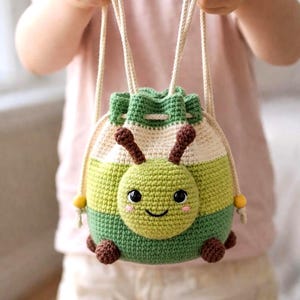 Puede incluir: Bolso con cordón hecho a mano en ganchillo, verde, blanco y beige, con una cara de oruga sonriente. El bolso tiene antenas marrones, pequeños pies marrones y cuentas amarillas en el cordón. El bolso es sostenido por una persona.