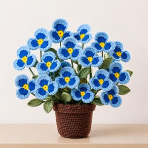 May include: A potted arrangement of crocheted flowers featuring three-petaled blossoms in shades of light and dark blue with bright yellow centers. The flowers are attached to green stems with textured green leaves, all housed in a brown crocheted pot.