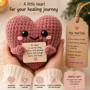 Op de afbeelding: Een oudroze gehaakt hart-knuffeltje met zwarte veiligheidsoogjes en roze wangen, dat een klein kaartje vasthoudt met de tekst: it's okay to not be okay. You're still growing. You're still enough. You're still loved. Inclusief een papieren label.