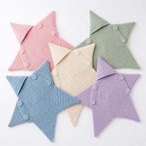 Può includere: Cinque cuscini all'uncinetto a forma di stella in colori pastello: rosa, blu, lavanda, crema e verde. Ogni cuscino ha una patta fissata con bottoni. I cuscini sono disposti su una superficie bianca.