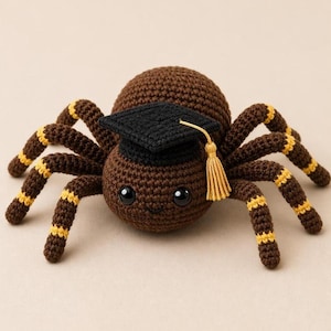 May include: A crocheted brown spider wearing a black graduation cap with a gold tassel. The spider has large, black eyes and a smiling expression. Its legs are brown with yellow stripes, and the spider is set against a neutral background.