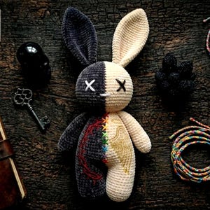 Puede incluir: Un conejo de peluche hecho a mano con un diseño dividido, con secciones negras y crema. El conejo tiene ojos bordados y un diseño cosido en el cuerpo. Las orejas son largas y el conejo está sobre una superficie de madera oscura.