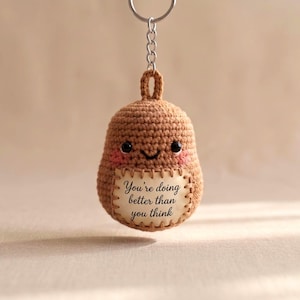 Puede incluir: Un llavero de ganchillo marrón hecho a mano con forma de patata sonriente con mejillas rosadas. El llavero tiene una pequeña etiqueta con el texto "You're doing better than you think". El llavero está unido a un llavero plateado.