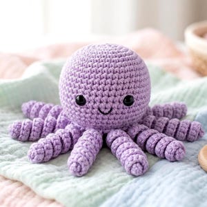 Puede incluir: Un juguete de pulpo de ganchillo lavanda hecho a mano con ojos de seguridad negros y una boca sonriente. El pulpo tiene ocho tentáculos rizados y descansa sobre una manta de colores pastel. Un anillo de dentición de madera está en primer plano.