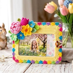 Könnte beinhalten: Ein handgefertigter gelber Häkel-Bilderrahmen, verziert mit bunten Häkelblumen. Der Rahmen enthält ein Foto einer lächelnden Person auf einer Wiese. Er hat einen Holzverschluss und eine Karte mit "SPRING JOY".