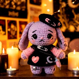 Puede incluir: Muñeca de conejo de ganchillo hecha a mano en tonos morado, rosa y negro. El conejo lleva una boina negra con un imperdible y una estrella, y un vestido negro con un corazón rosa, una luna y un dije de calavera. La muñeca tiene ojos negros grandes.