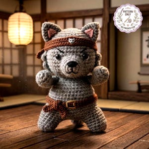 May include: A crocheted gray wolf doll in a martial arts pose, wearing a brown headband and belt. The doll has pink inner ears and a stern expression. The image includes a circular sticker that says "PATTERN ONLY NO FINISHED ITEM".
