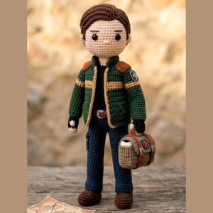 Könnte beinhalten: Handgefertigte Häkelpuppe einer Person in einer grünen Jacke mit braunen Akzenten, blauen Jeans und braunen Stiefeln. Die Puppe hat braunes Haar und hält eine kleine braune Tasche. Sie steht auf einer Holzoberfläche.