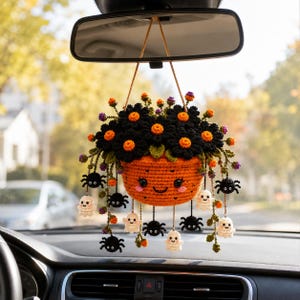 May include: A crocheted car mirror hanging ornament featuring an orange pumpkin-shaped pot with a smiling face, filled with black flowers with orange centers, and dangling small white ghosts and black spiders on thin brown strings.