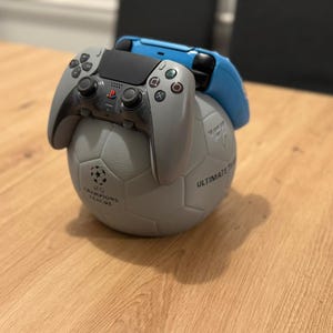 May include: A gray and black PlayStation 5 controller rests on a gray soccer ball-shaped stand, with a blue controller behind it. The soccer ball has the text "ULTIMATE TEAM" and a logo. The items are on a wooden surface.