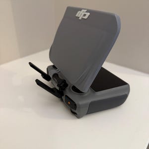 Puede incluir: Un control remoto de dron gris con pantalla y antenas. El control remoto tiene una cubierta protectora con el logotipo de DJI. El botón de encendido está iluminado en naranja. El dispositivo está sobre una superficie blanca.