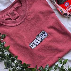 Op de afbeelding: Een stoffig roze t-shirt met het woord "RISK" geborduurd in een Scrabble-achtig ontwerp. Een speelgoed raceauto is zichtbaar op de achtergrond. Het shirt is gemaakt van een zacht materiaal en heeft een ronde hals.
