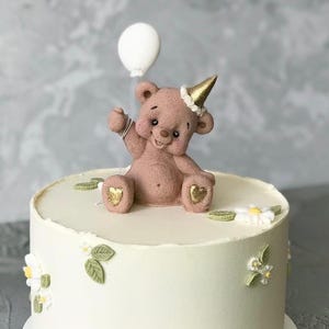 May include: A round, white frosted cake decorated with white flowers, green leaves, and a pink teddy bear wearing a gold party hat. The bear holds a white balloon. The cake is on a white cake board.