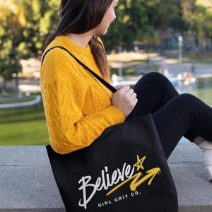 Könnte beinhalten: Eine schwarze Tragetasche mit dem weißen Schriftzug Believe, einem gelben Stern und einer gelben Pfeilgrafik sowie dem kleineren weißen Text GIRL GRIT CO. unter dem Hauptwort. Die Tasche verfügt über schwarze Schulterriemen.