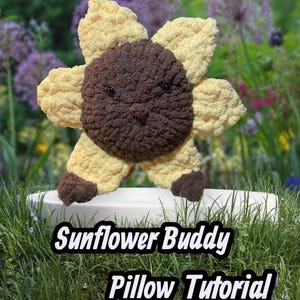 May include: A crocheted sunflower pillow in yellow and brown, with a smiling face. The pillow is placed on a white surface against a backdrop of greenery and flowers. The text "Sunflower Buddy Pillow Tutorial" is displayed at the bottom of the image.