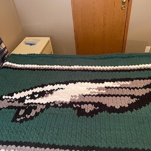 May include: A large, hand-knitted blanket in shades of green, black, white, and gray. The blanket features a stylized eagle design in the center. The blanket is made of thick, chunky yarn.