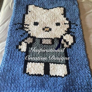 Puede incluir: Una manta azul hecha a mano con una imagen grande y pixelada de un gato blanco con detalles en negro. El gato lleva un atuendo gris. La manta tiene el texto "Inspirational Creation Designs" en escritura blanca.