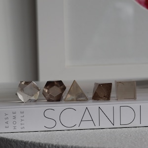 May include: Five geometric crystal shapes in varying shades of brown and clear, arranged on a white book with the word "SCANDI" printed on it. The shapes include a cube, a pyramid, and a multi-faceted design.