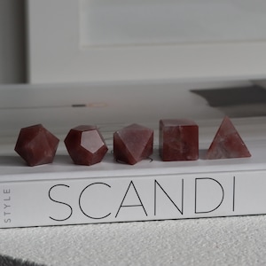 May include: Five geometric rose quartz crystals in various shapes: a rhombicuboctahedron, a dodecahedron, a truncated octahedron, a cube, and a triangular pyramid. The crystals are arranged on a white book with the word "SCANDI" printed on it.