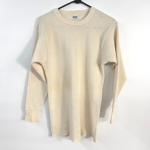 May include: A long-sleeve thermal shirt in an off-white cream colour, featuring a textured waffle-knit pattern, a crew neckline, and ribbed cuffs at the wrists. The garment is displayed on a black hanger against a plain white background.