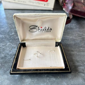 Puede incluir: Un joyero vintage con interior color crema y marco exterior negro. El interior de la tapa presenta el texto Shields Fifth Avenue en letra negra. La base contiene tres pequeños soportes de alambre metálico en tono plateado.