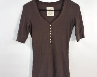 May include: A dark brown ribbed henley shirt with three-quarter length sleeves, a deep scoop neckline featuring a ruffled trim, and a front placket with seven small white buttons. A small white embroidered bird logo is visible at the bottom hem.