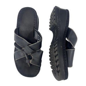 May include: A pair of black sandals featuring a toe-post design with crisscrossing straps accented by white contrast stitching. The sandals have a thick, molded black sole with a textured tread pattern on the bottom for grip and stability.