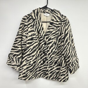 Puede incluir: Una chaqueta de manga larga con estampado de cebra en blanco y negro. La prenda incluye solapas de muesca, tres botones negros en la parte delantera y dos bolsillos con solapa. El interior está forrado con tela beige lisa.
