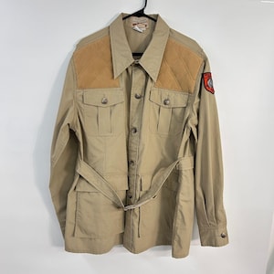 May include: A tan utility jacket featuring quilted brown shoulder patches, a button-front closure, and a matching waist belt. The garment includes four front pockets with buttoned flaps and a red and black circular patch on the left sleeve.