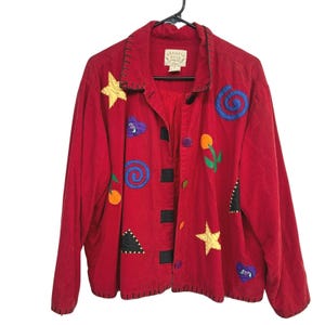 May include: A red long-sleeve jacket featuring a collar with black whipstitch detailing. The garment is decorated with various colorful fabric appliqués including yellow stars, blue spirals, orange flowers, and purple hearts. It has a button-front closure.