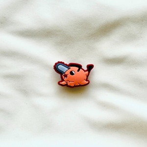May include: A shoe charm featuring a cartoon character with an orange body, a red outline, and a blue chainsaw. The charm has a smiling face and black details. The background is a white fabric.