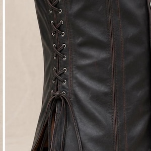 May include: A dark brown leather corset featuring vertical stitched panels and a side lacing system with silver-toned metal eyelets. The garment includes long, thin leather laces tied in a knot at the bottom, creating a structured fit.