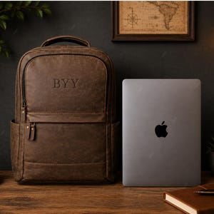 May include: A brown leather backpack featuring the embossed letters BYY on the front pocket, accompanied by a closed silver laptop positioned to its right. The backpack includes a top handle, a front zippered compartment, and side pockets.