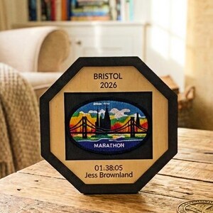 Könnte beinhalten: Achteckiger Holzrahmen mit einem Bild des Bristol 2026 Marathon mit Brücke und Skyline. Der Rahmen steht auf einem Holztisch mit einer Medaille, einer Startnummer und Laufschuhen. Der Text "01:38:05 Jess Brownland" ist sichtbar.