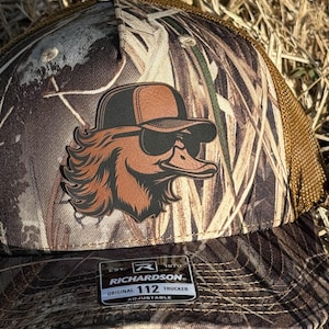 May include: Camouflage trucker hat with a brown and black duck graphic wearing sunglasses. The hat has a mesh back and a Richardson 112 Trucker label.