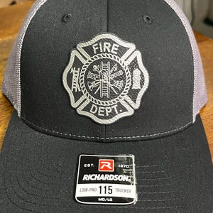 May include: Black and gray trucker hat with a fire department emblem. The hat features a black front panel and brim, a gray mesh back, and a silver emblem with the words "FIRE DEPT." The hat has a Richardson tag.