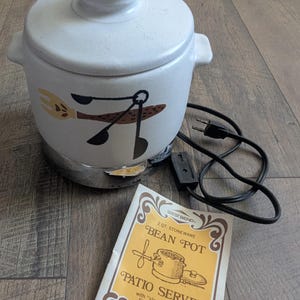 Puede incluir: Una olla de frijoles West Bend vintage con tapa, con un acabado blanco moteado y un diseño decorativo de utensilios de cocina. La olla se asienta sobre una base cromada e incluye un cable de alimentación y una etiqueta "Patio Server".