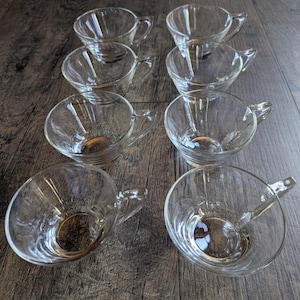 May include: A set of eight clear glass teacups arranged in two columns of four. Each cup features a smooth, rounded bowl shape with a small, integrated handle on the side. The glass is transparent, showing a simple, functional design for hot beverages.