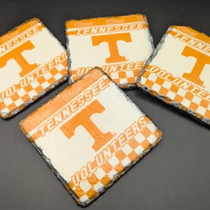 May include: Four square slate coasters with the University of Tennessee Volunteers logo. Each coaster features the orange "T" logo on a white background, with the words "Tennessee Volunteers" and a checkered orange and white border.