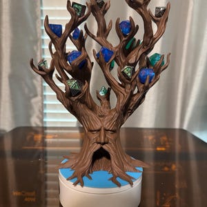 Puede incluir: Un soporte para dados en forma de &aacute;rbol marr&oacute;n con una cara tallada en el tronco. El &aacute;rbol contiene varios dados de m&uacute;ltiples caras en varios colores, incluyendo azul, verde y plateado. La base es blanca con un acento azul.