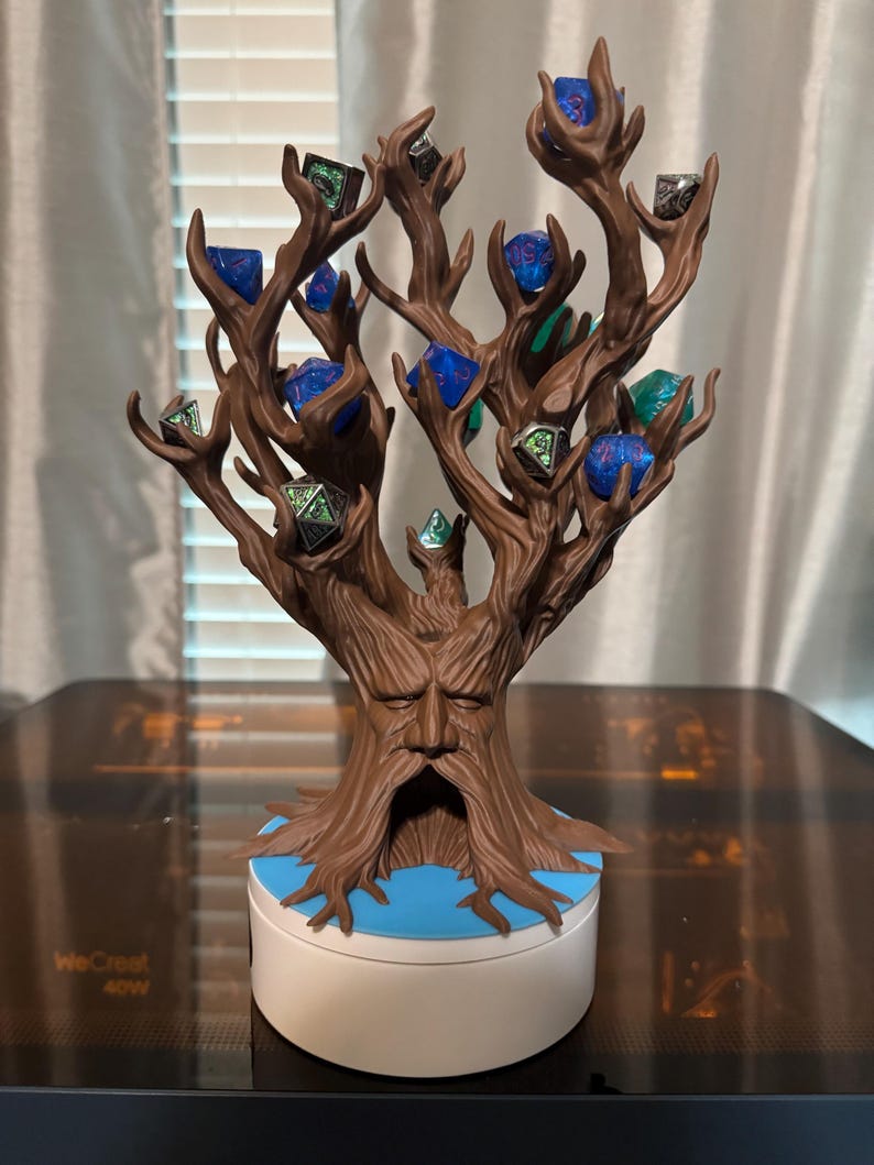 Puede incluir: Un soporte para dados en forma de &aacute;rbol marr&oacute;n con una cara tallada en el tronco. El &aacute;rbol contiene varios dados de m&uacute;ltiples caras en varios colores, incluyendo azul, verde y plateado. La base es blanca con un acento azul.