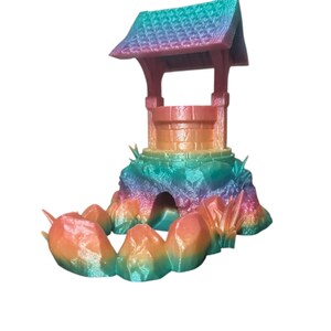 May include: A colorful 3D-printed wishing well model. The well features a roof, posts, and a brick-like structure, all in a gradient of rainbow colors. The well sits on a rocky base with grass-like details, also in a rainbow gradient.