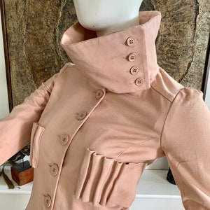 May include: A blush pink collared jacket with a high neckline and multiple buttons. The jacket has a pleated detail on the front and long sleeves.