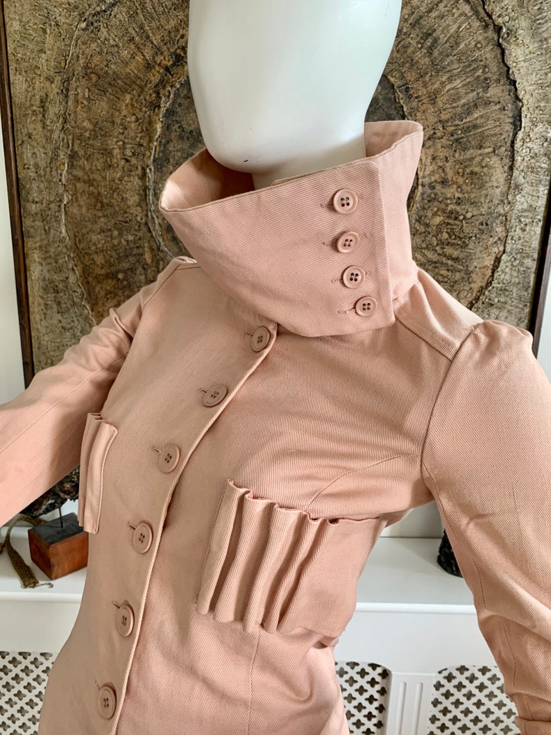 May include: A blush pink collared jacket with a high neckline and multiple buttons. The jacket has a pleated detail on the front and long sleeves.