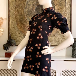 May include: A black mini dress with a high neck and short sleeves. The dress has a floral print in shades of yellow, orange, and burgundy.