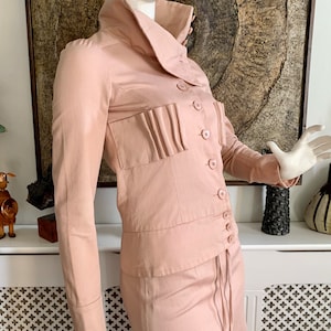 May include: A blush pink collared jacket and skirt set with a long skirt and a button-up front. The jacket has a large collar and a pleated detail on the front.