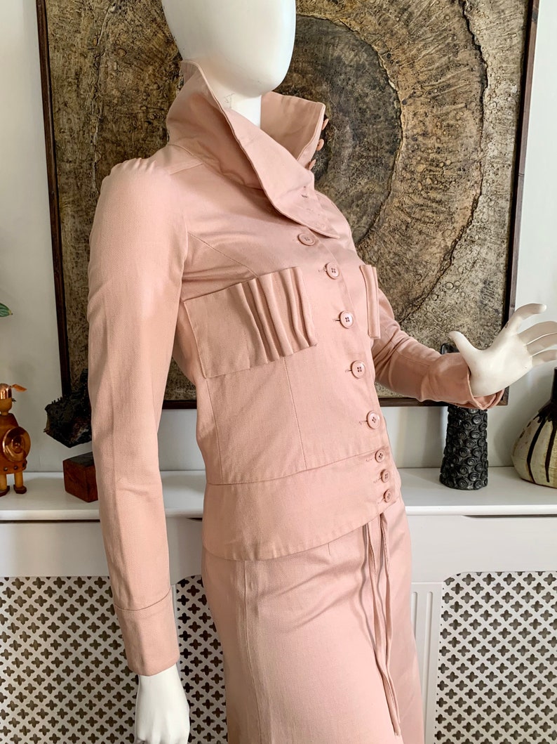 May include: A blush pink collared jacket and skirt set with a long skirt and a button-up front. The jacket has a large collar and a pleated detail on the front.