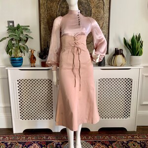 May include: A blush pink satin blouse with a high neck and button closure. The blouse has long sleeves with a gathered puff at the shoulder. The blouse is paired with a blush pink skirt with a lace-up detail at the waist.