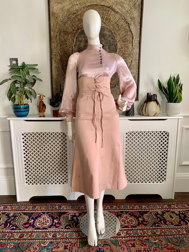 May include: A blush pink satin blouse with a high neck and button closure. The blouse has long sleeves with a gathered puff at the shoulder. The blouse is paired with a blush pink skirt with a lace-up detail at the waist.