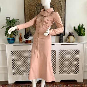 May include: A blush pink skirt suit with a long skirt and a fitted jacket featuring a high collar and a button closure. The jacket has a pleated detail on the front.